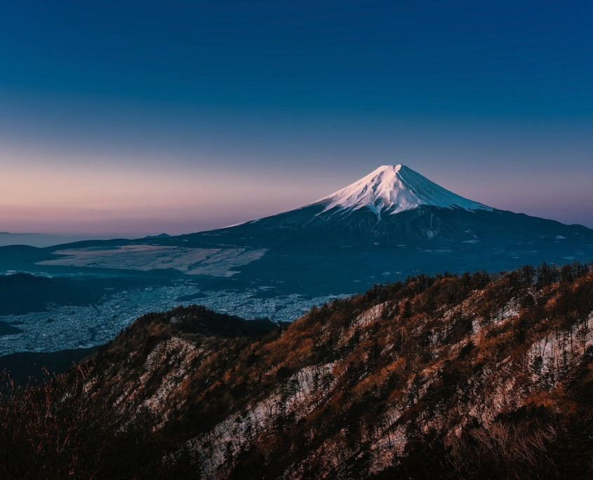 kanenori-mount-fuji-6874364_1280_scaled Mount Fuji's snow-covered cone at dusk, seen from a forested ridge above the valley